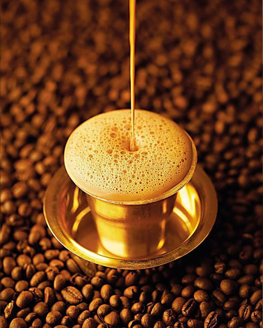Pouring frothy South Indian filter coffee into a traditional brass dabara set resting on roasted coffee beans.
